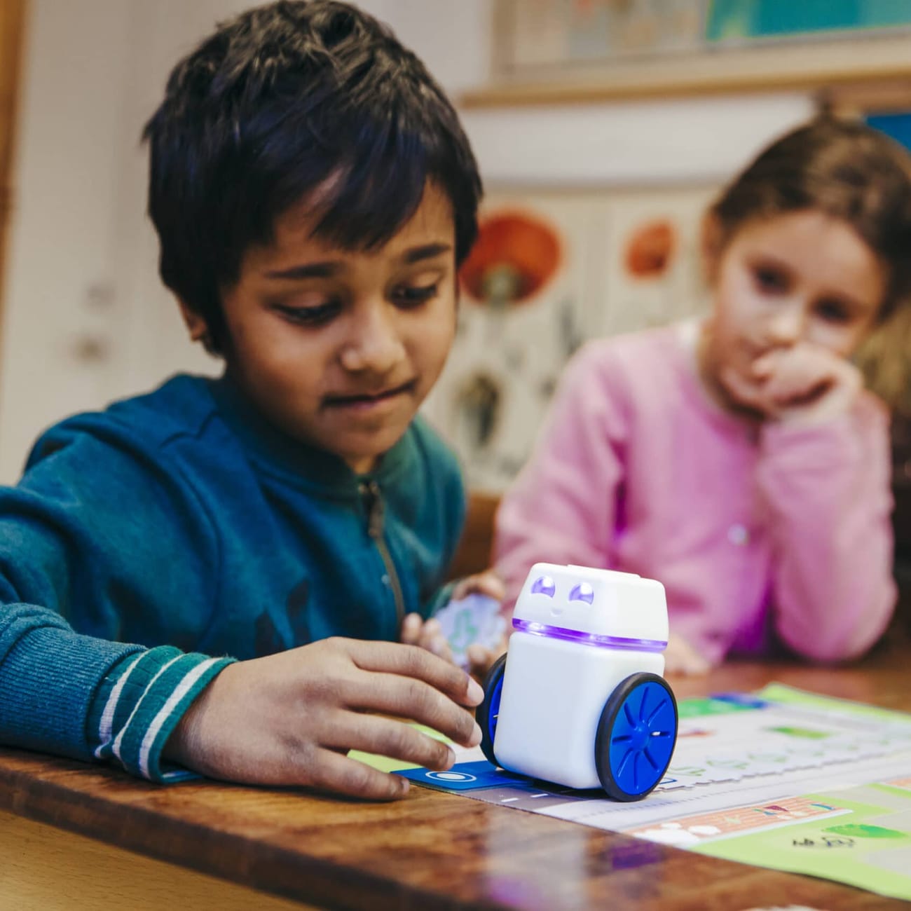 A student at a classroom desk with a hand on the KUBO robot as it sits on a row of TagTiles, a classmate watching beside it